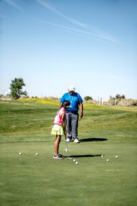 A girl practicing golf under sunny skies with her coach on a green course.