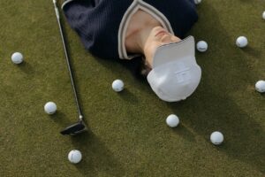 Asian woman in white cap lying on golf course with golf balls and club.